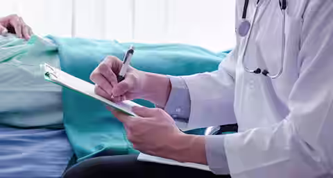 A doctor wearing a white coat and a stethoscope writes on a clipboard while sitting beside a bed. A patient is lying in the bed, covered with a turquoise blanket. The setting appears to be a hospital or medical facility.