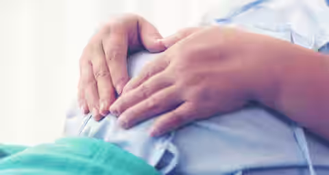 A close-up of a person wearing a light blue hospital gown, lying down and resting their hands on a pregnant belly. The image focuses on the hands and the belly, with soft lighting in the background.