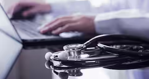 A stethoscope rests on a glossy black surface in the foreground, while a person wearing a white coat types on a laptop in the background. The image suggests a medical professional working on digital documentation.
