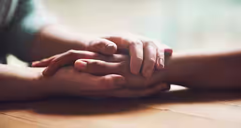 Close-up of one person gently holding another person's hand on a wooden table, conveying comfort, support, or empathy in a warm, soft-lit setting.