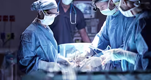 A surgical team in an operating room performs a procedure. Four medical professionals, dressed in blue scrubs and sterile gloves, are focused on the patient. The environment is sterile, and surgical instruments and monitors are visible in the background.
