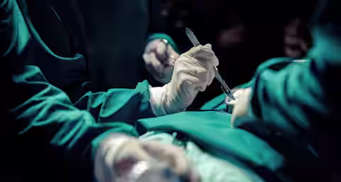 Surgeons in green scrubs and white gloves performing a surgical procedure. One surgeon holds a scalpel, and another assists. The scene is well-lit with focus on the patient's torso, while the background remains dark and out of focus.