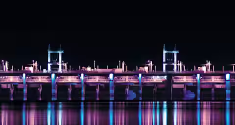 A brightly lit dam at night reflecting on the calm water below. The structure is illuminated with blue and purple lights, creating a vivid and serene scene.