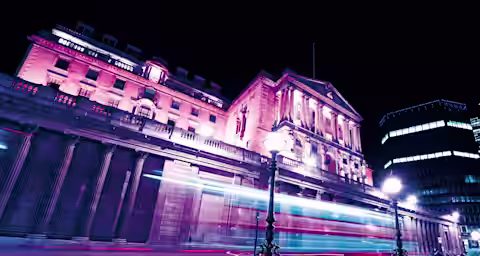 A historic building illuminated with pink and purple lights at night. Long exposure creates light trails from passing vehicles, enhancing the dynamic scene. Street lamps and modern buildings in the background contrast with the classical architecture.