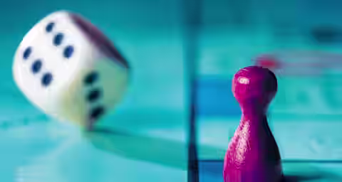 A close-up image of a red game piece and a tumbling white die with black dots on a blue game board. The die is shown mid-roll, indicating movement, while the game piece stands stationary. The game board has a grid pattern.