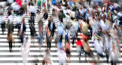 A blurred image of numerous people walking across a large, busy, zebra-striped crosswalk. The motion blur captures the hustle and bustle of the crowd, giving a sense of fast-paced urban life. The scene is vibrant with diverse clothing colors and patterns.