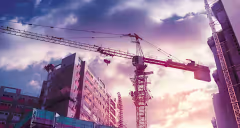 A construction site with tall cranes and buildings under construction. The sky is filled with dramatic clouds illuminated by a colorful sunset, casting a warm glow on the scene. Scaffolding and partially completed structures are prominent in the foreground.