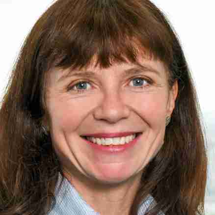 A woman with shoulder-length brown hair is smiling at the camera. She is wearing a dark blazer over a light blue collared shirt. The background is a blurred outdoor scene, possibly showing buildings and greenery.