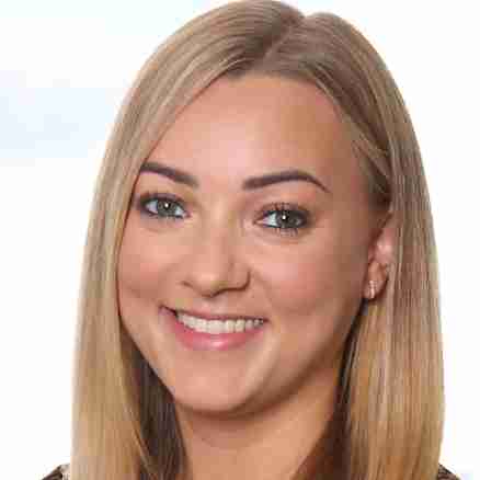 A woman with long, straight blonde hair is smiling at the camera. She is wearing a black dress with a small orange and white pattern. The blurred background indicates she is indoors with a window showing a cityscape behind her.