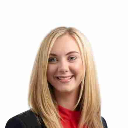 A woman with long blonde hair is smiling and wearing a black blazer over a red top. The background is white.