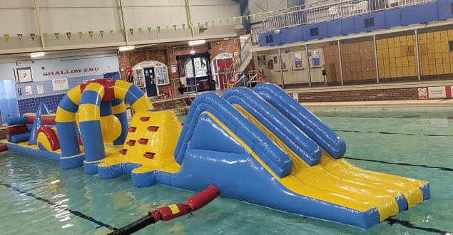 Inflatable water obstacle course in a pool, featuring slides, climbing wall, and arches. It's colourful, mainly yellow and blue, with a fun, playful vibe.