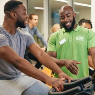 The image shows a fitness instrcutor showing a mae gym member how to use an exercise bike.