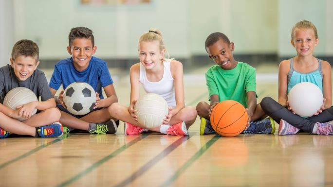 A group of children sitting cross legged holding various sports balls