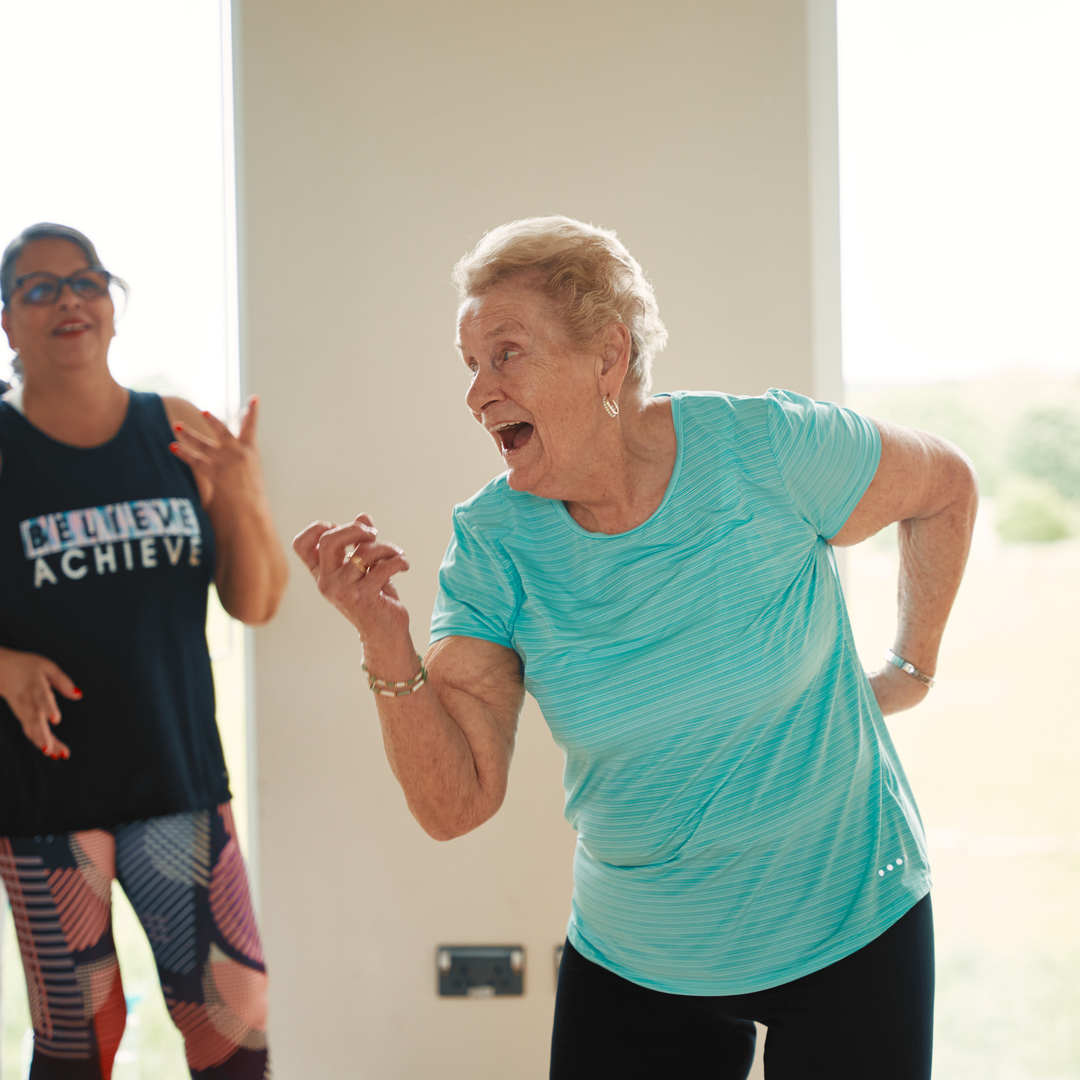 image shows a senior lady in a dance pose with a big smile on her face as others watch on smiling.