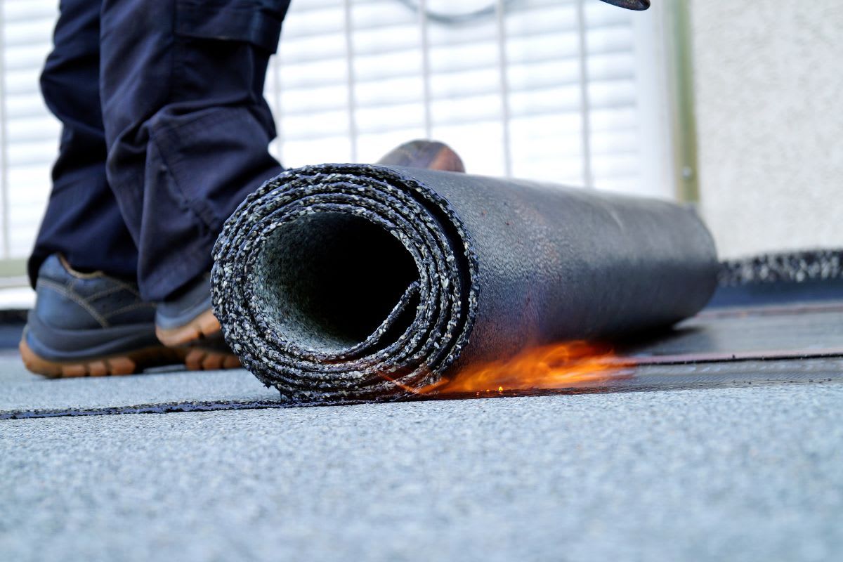 Arbeiter rollt eine neue Bitumenbahn mit einem Gasbrenner auf einem Flachdach aus, wobei Flammen am Rand der Rolle zu sehen sind.