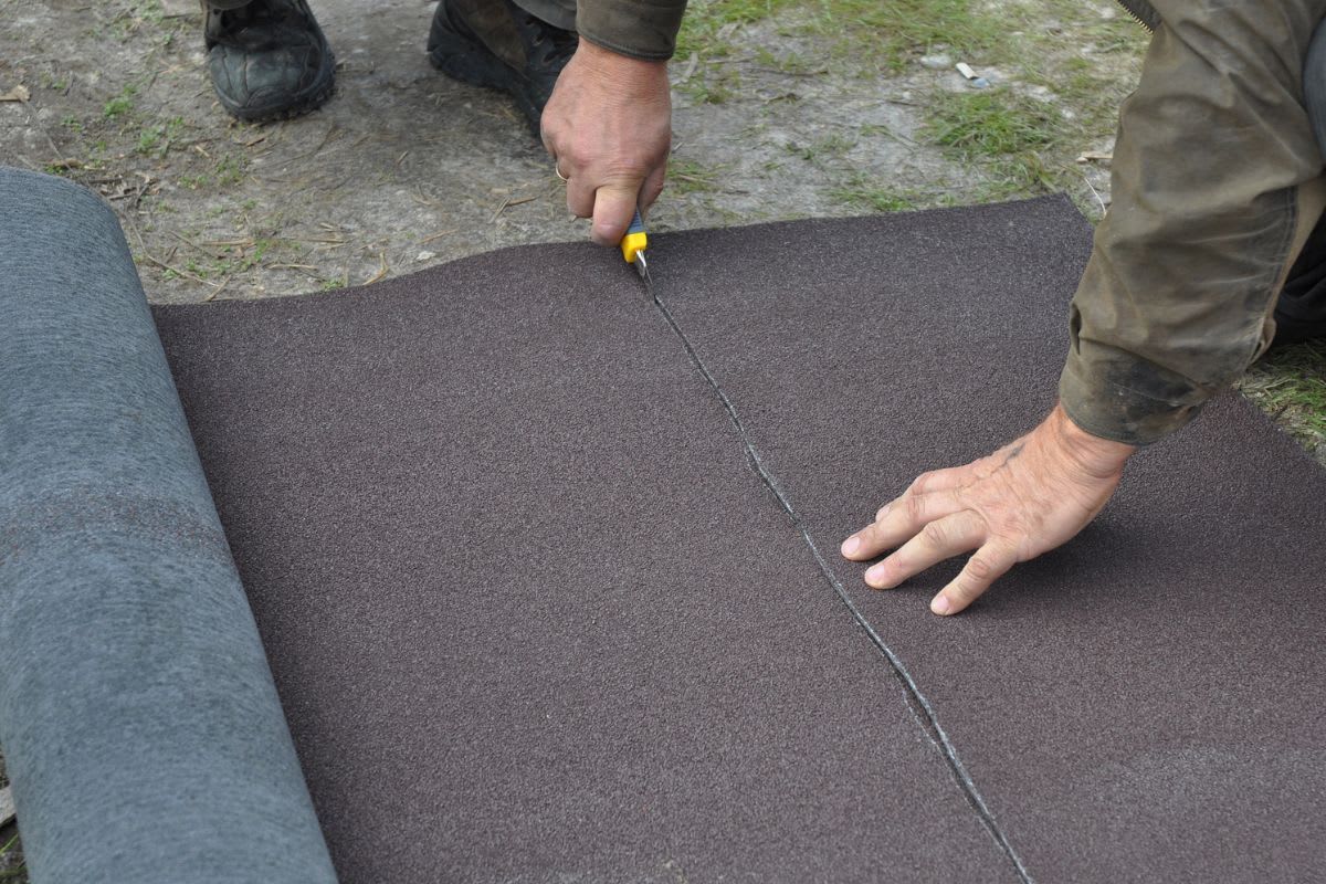 Person schneidet Dachpappe mit einem Cutter auf einer Unterlage zu, während sie die Pappe mit der anderen Hand festhält.