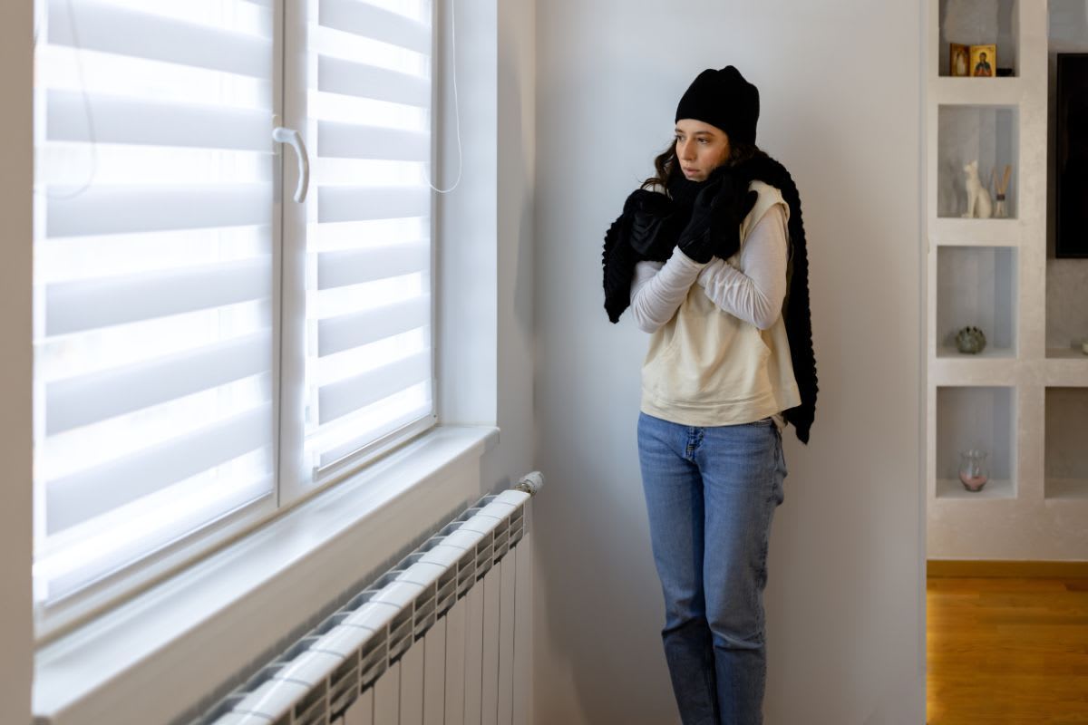 Person in Winterkleidung friert in einem Raum neben einem Heizkörper, was auf ein möglicherweise defektes Thermostat hinweist.