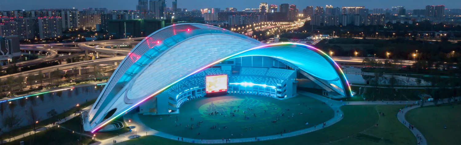 Modern architectural pavilion with a curved, illuminated white roof and a large outdoor screen at dusk.