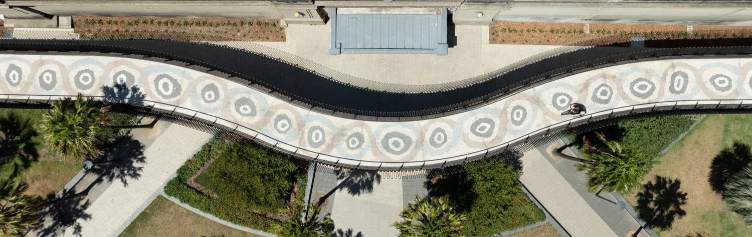 Aerial view of a curved pedestrian walkway with circular patterns next to a train track and palm trees.