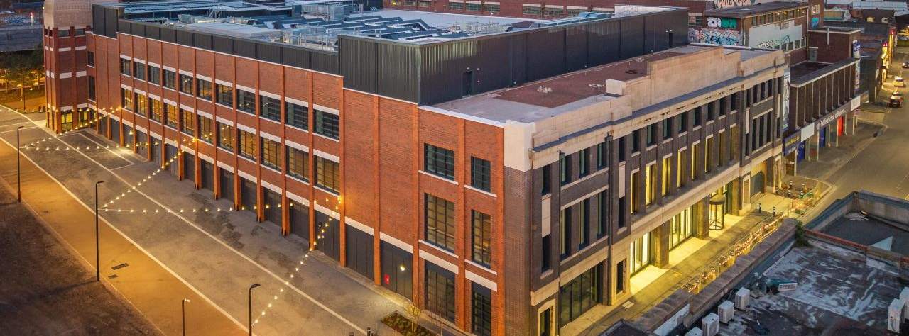 A multi-story red brick commercial building at dusk with interior lights on and a city skyline in the background.