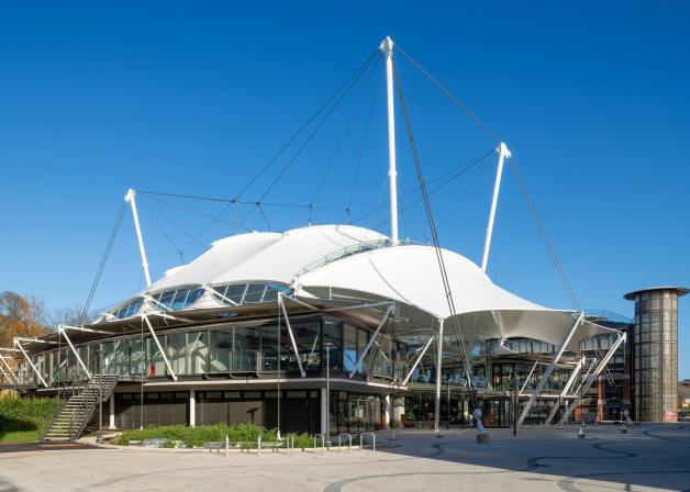 A modern building with a large, white tensile membrane roof supported by tall masts and steel cables.