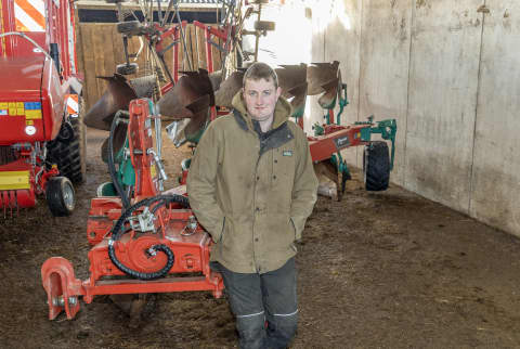 Tom Wayne, wearing a brown jacket, standing in front of his red and green Kverneland 2300 S Reversible Plough in his barn