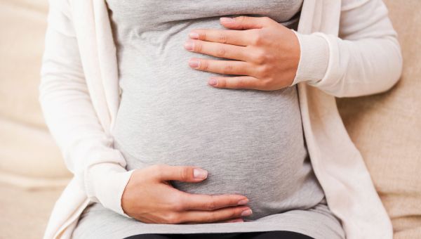 pregnant person dressed in a light gray shirt and cream colored sweater, sitting down and holding their belly