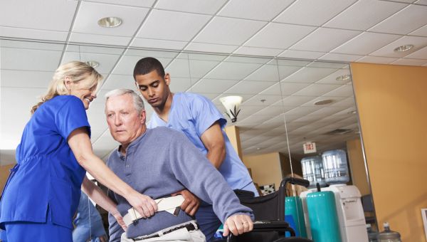 a male and female nurse lift an elderly man out of a wheelchair