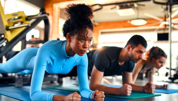 A Black woman in a turquoise workout suit does a plank exercise in a gym. In the background are several men also doing planks.