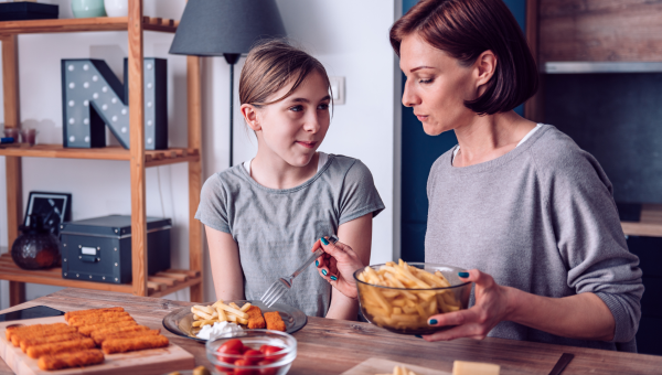 preteen girl with autism and mother sit at the dining table serving a large portion of fish sticks and fries and a small portion of yogurt