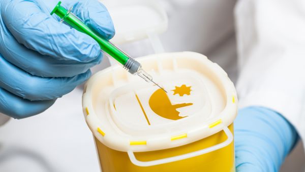 gloved hand of a medical professional disposes of a syringe in a medical waste bucket