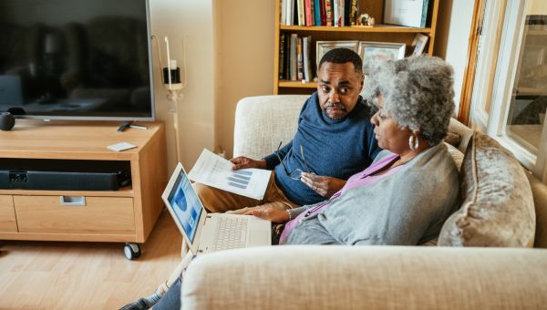 an elderly Black man and woman sit in their living room on a couch, the man reading a magazine and looking at his wife, who is working on a laptop computer 