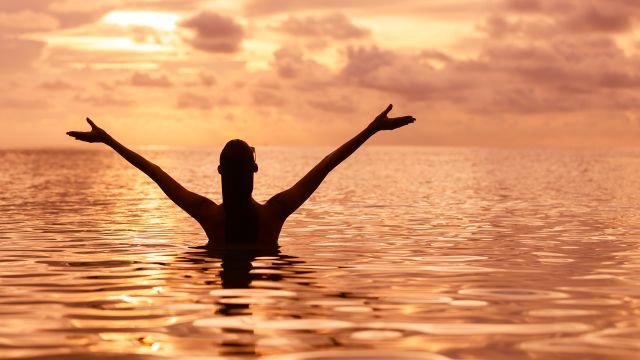 person in body of water with arms raised at sunset