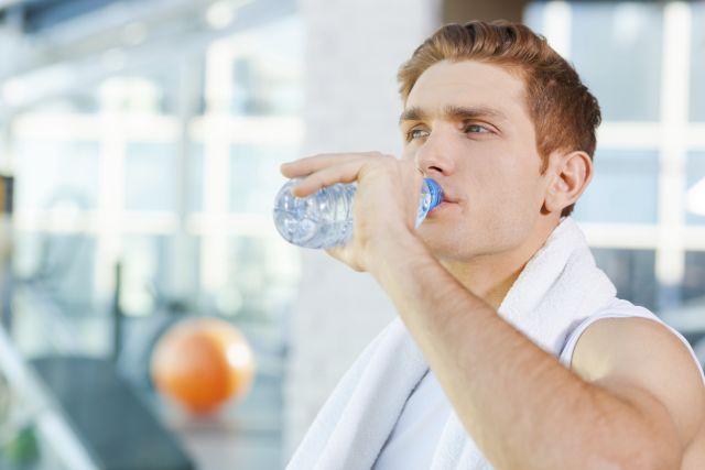 Tired young man carrying towel on shoulders and drinking water while standing in gym