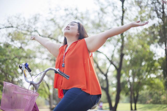 A woman rides a bike and throws her arms up in the air with happiness as she exercises.