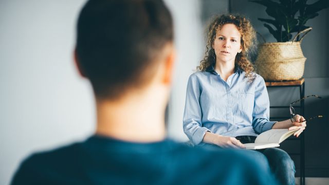 Female therapist holds notebook and chats with male patient in an office. 