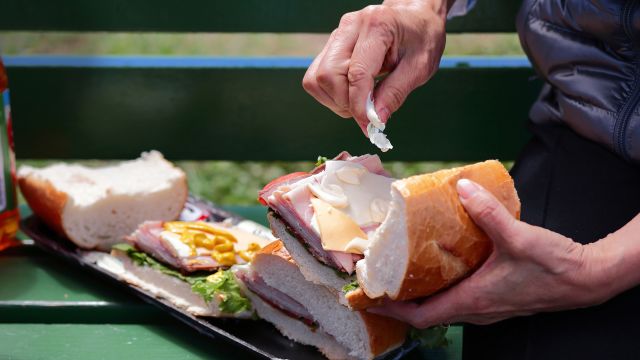 Man putting mayonnaise on a sandwich on a park bench.