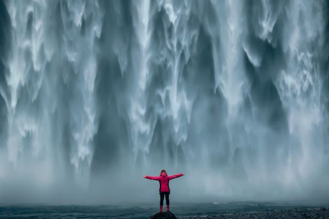 a woman stands with arms outstretched in front of a massive waterfall
