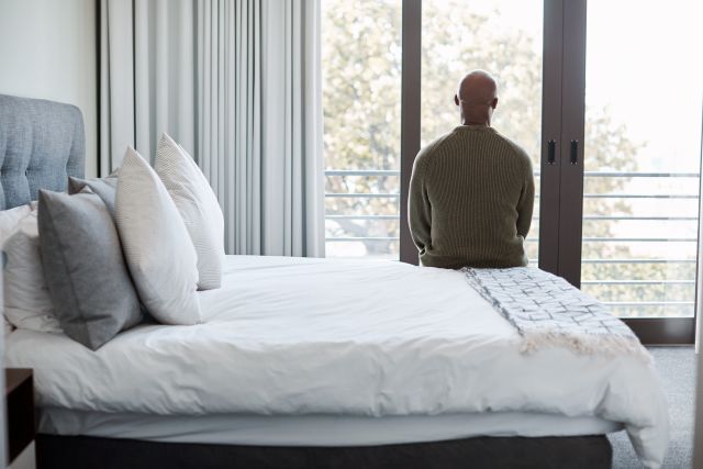 a Black man stares out the window of his bedroom, his back to the camera