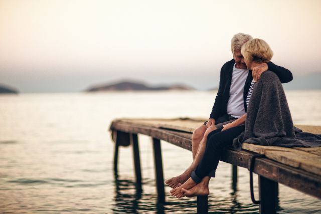 a middle aged man and woman sit in an embrace on a dock overlooking a lake