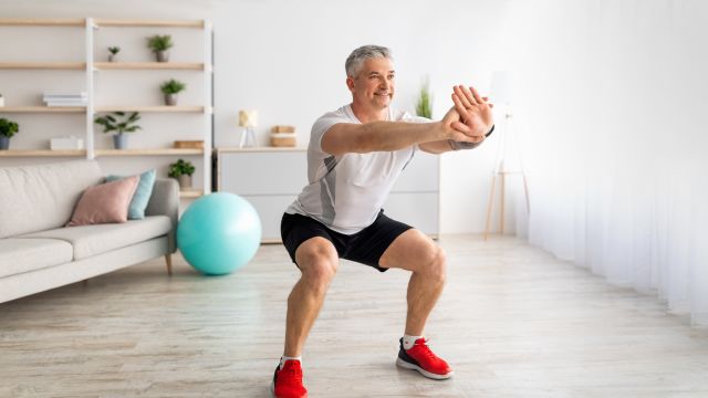 A smiling middle aged white man performs a body weight squat while exercising at home 