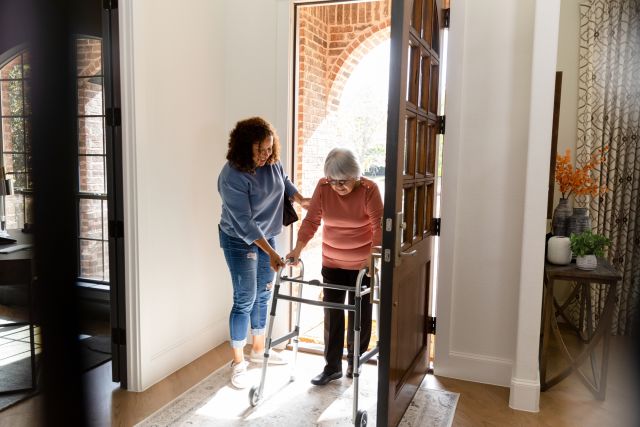 Woman helps her elderly mother with walker through the door of her home