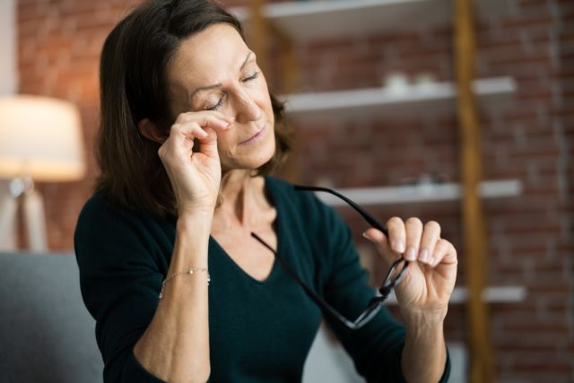 Woman at home holds her eyeglasses and rubs her irritated eyes 