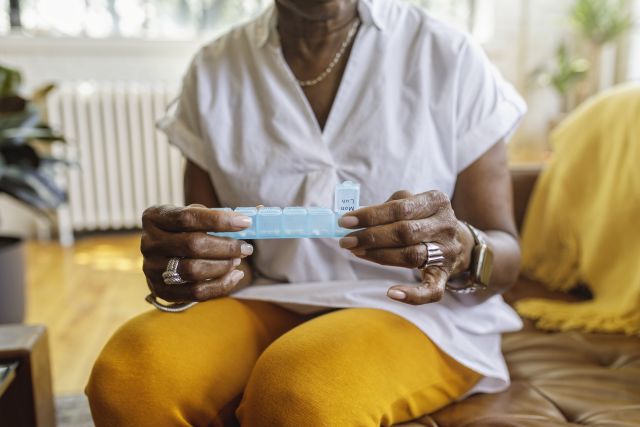 Senior black woman sits on the couch at home and takes medications from a daily pill organizer. 