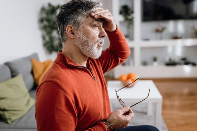 Sweaty mature man with worried facial expression sitting on living room couch holding hand on his forehead