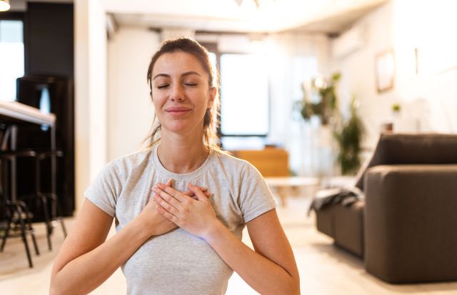 Young woman sitting on the floor meditating at home, holding her hands over her heart with eyes closed