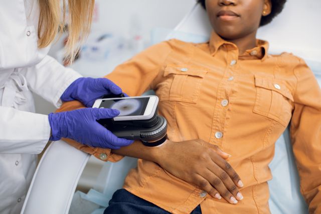 Close up of hands of female doctor dermatologist holding new generation dermatoscope, examining birthmarks and moles of a woman lying on her back on an exam table