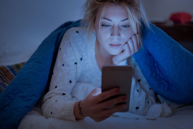 young woman in her bed in the dark under a blanket looking at her phone