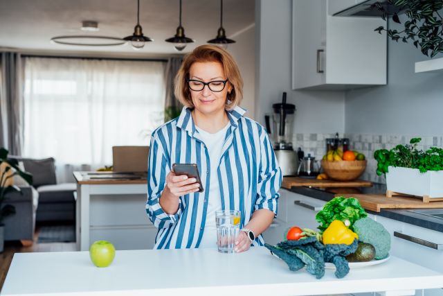 Woman in her kitchen holding a glass of lemon water, standing near a plate of vegetables, looking at her phone for meal planning