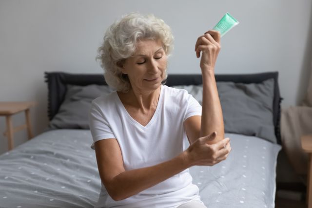 senior woman with eczema sitting on a bed putting a daily moisturizer on her arms